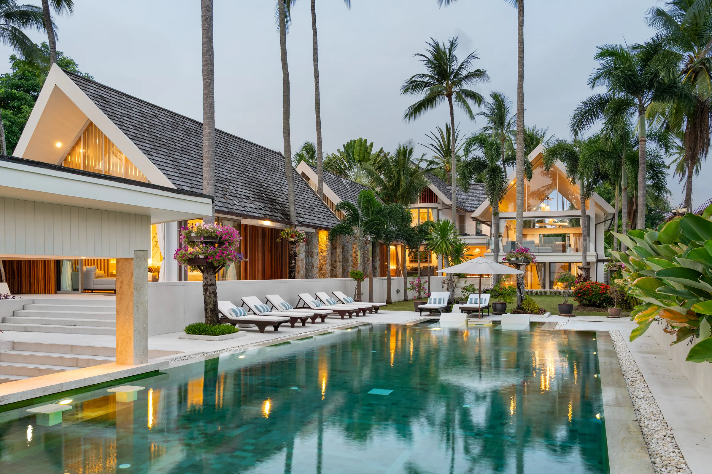 A villa pool at dusk with pavilions lit beyond it